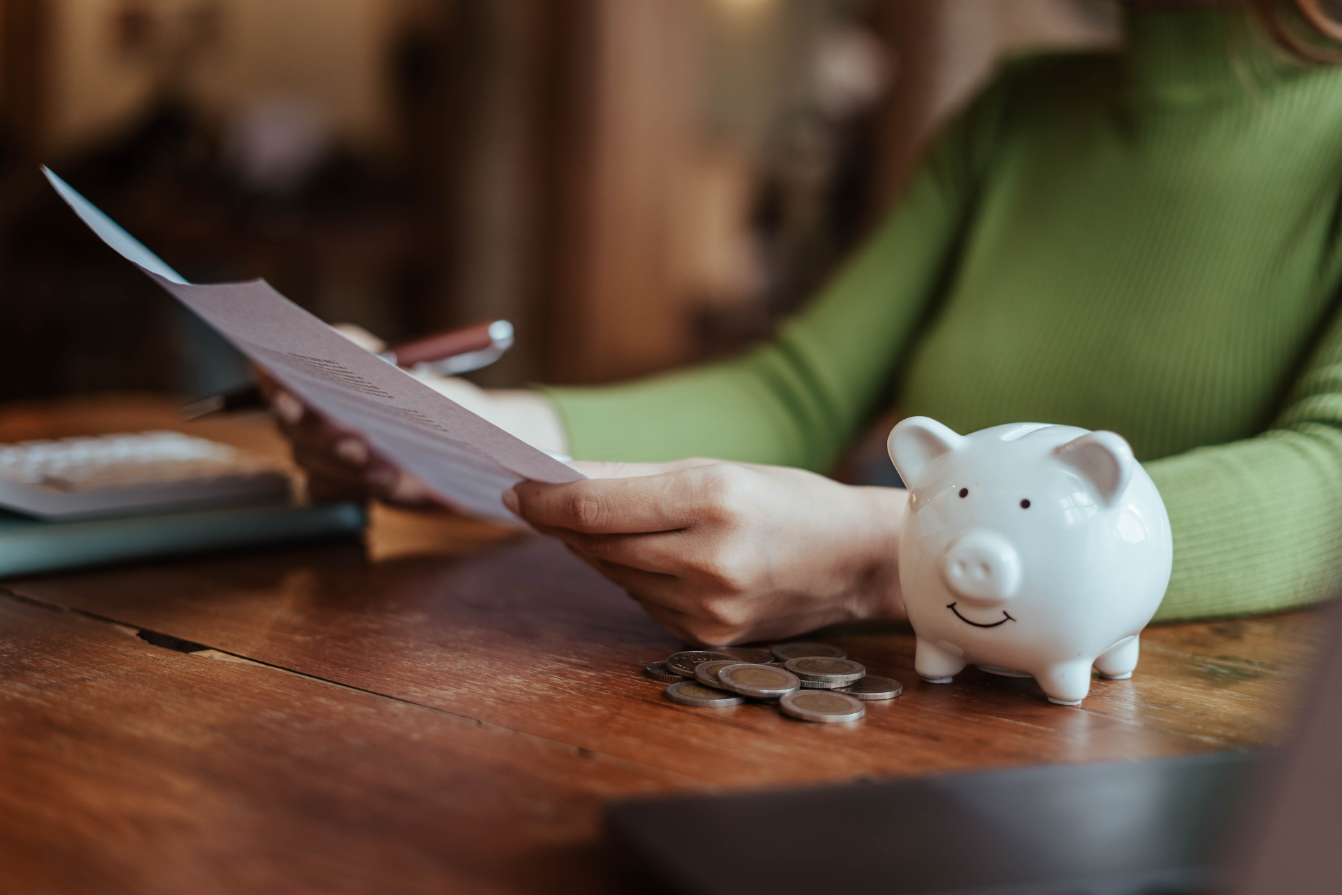 A white piggy bank on a wooden desk next to scattered coins, with a person reviewing financial documents in the background.